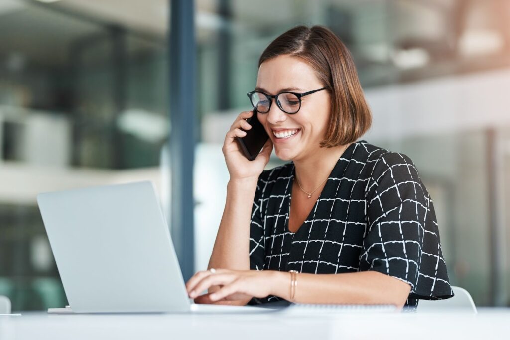 A smiling PT owner sits at a white desk in a bright, modern office. She is talking on a smartphone while simultaneously typing on a laptop.