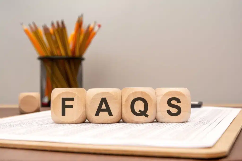 Four wooden blocks spelling "FAQS" sit on a clipboard with a spreadsheet, with a cup of pencils in the blurred background.