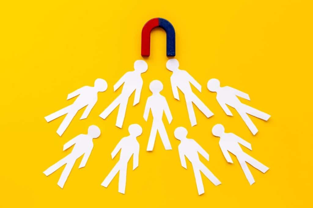 A lead magnet is attracting several white paper cutout figures toward it on a bright yellow background.