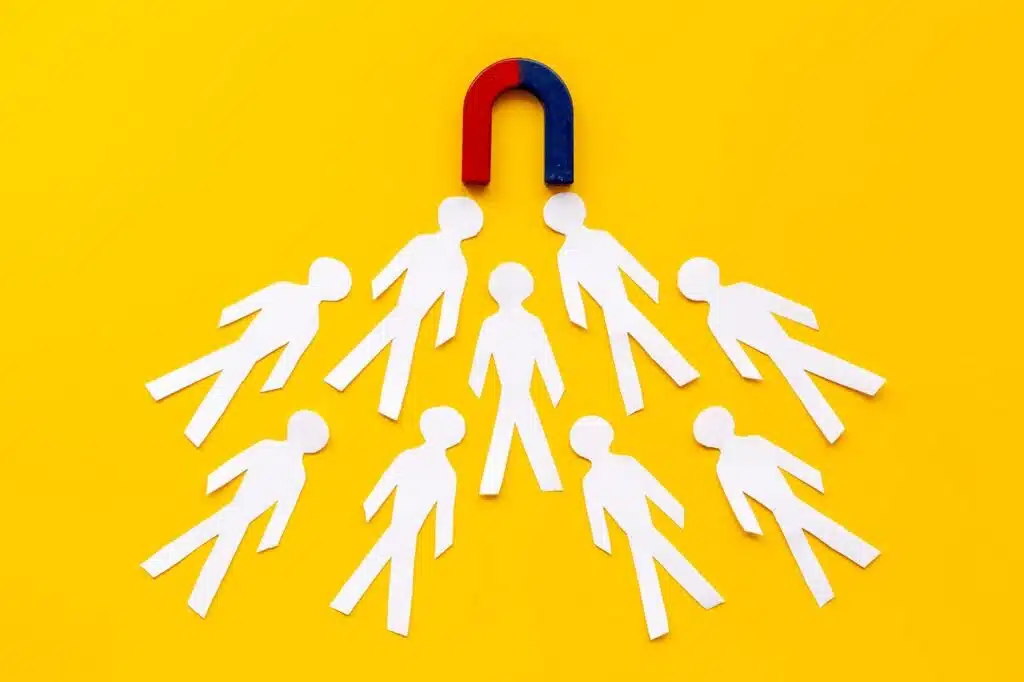 A lead magnet is attracting several white paper cutout figures toward it on a bright yellow background.