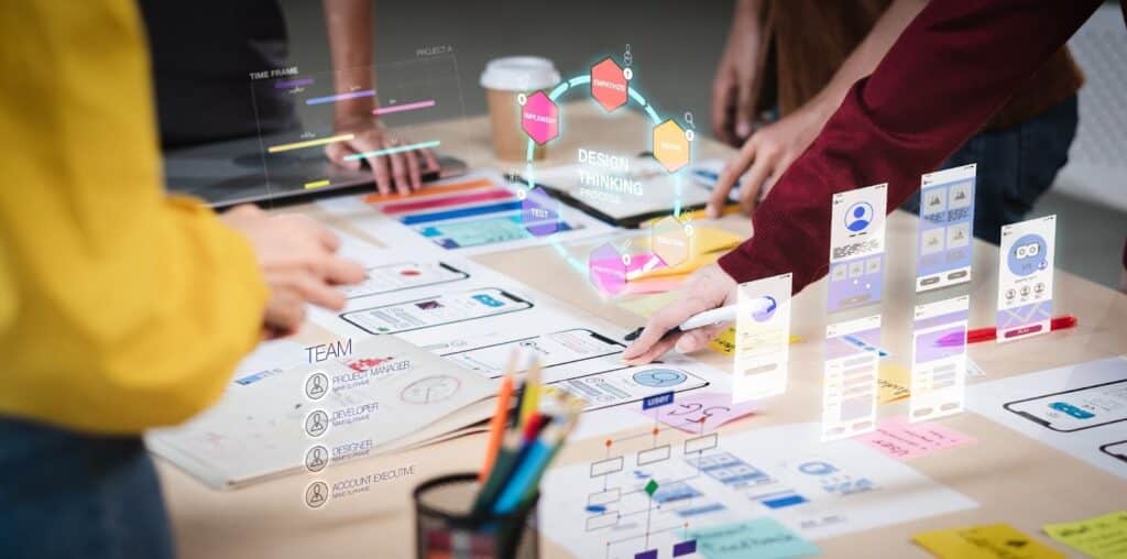 A team of four people works together on a desk covered with mobile app UI/UX designs, flow charts, and a visible "Design Thinking Process" diagram overlay.