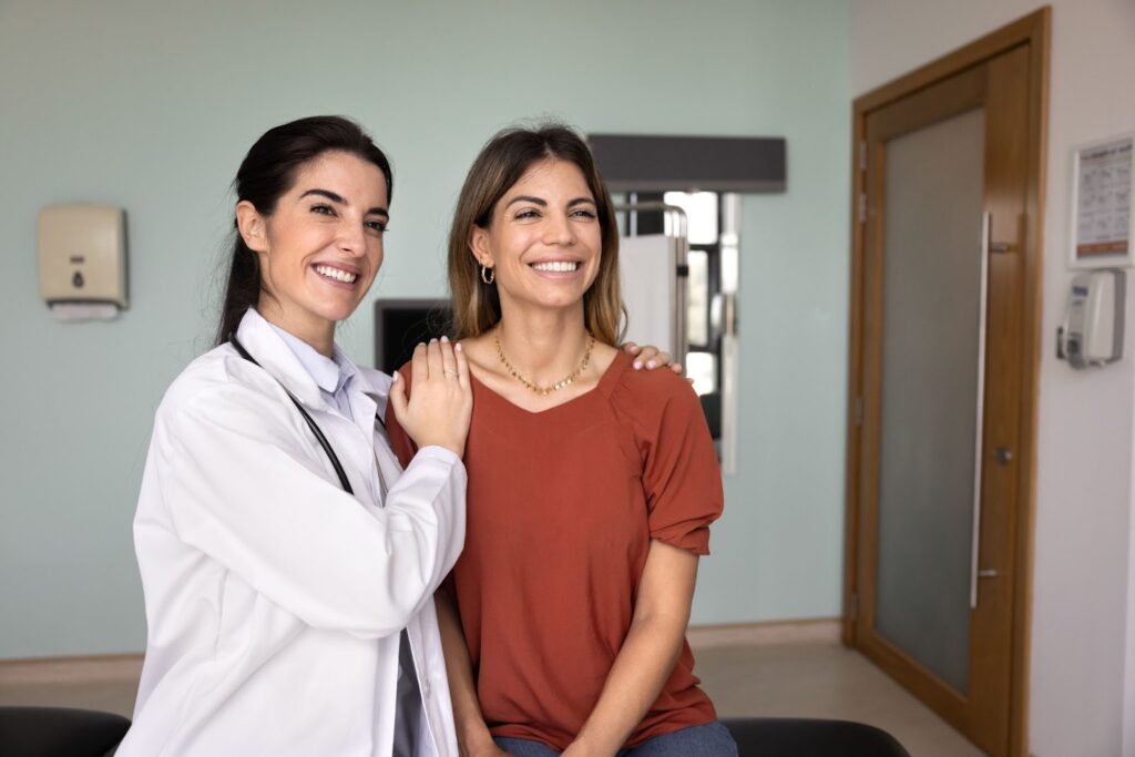 A female doctor in a white coat stands behind a female patient, both smiling and looking to the side.