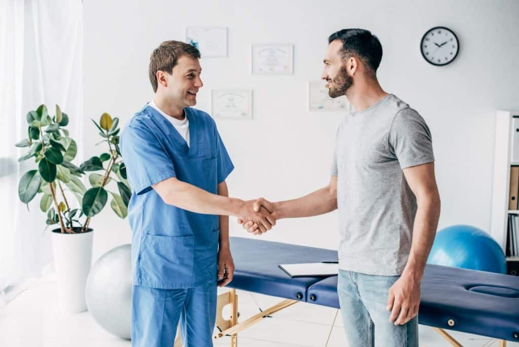 A male physical therapist in blue scrubs shakes hands with a male patient in a grey shirt and jeans in a brightly lit clinic room.