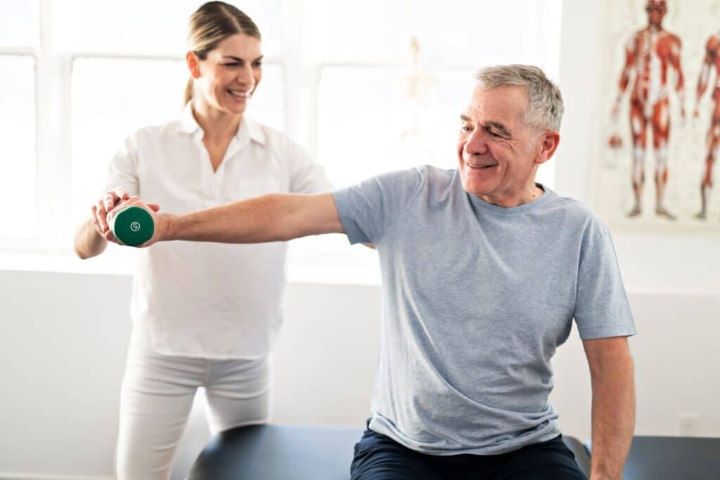 A physical therapist, smiling, assists an older man with a shoulder exercise using a small green dumbbell.