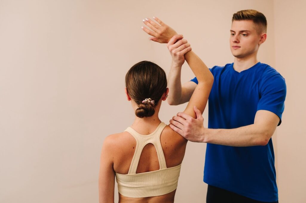 A physical therapist in a blue t-shirt assists a female patient with shoulder mobility exercises.