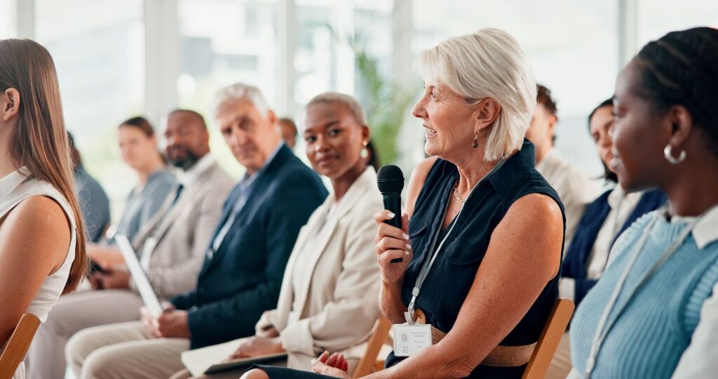 An older woman with short blonde hair speaks into a handheld microphone while sitting in the audience.