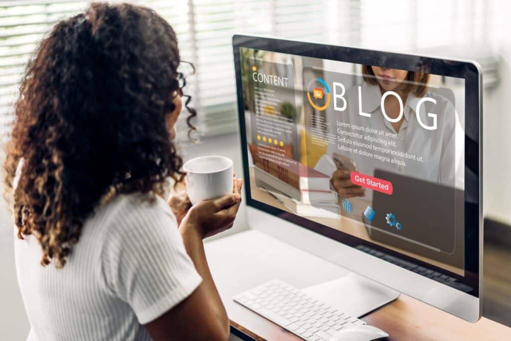 A woman with curly hair is sitting at a desk, looking at a computer monitor displaying a "BLOG" website, while holding a white coffee mug.