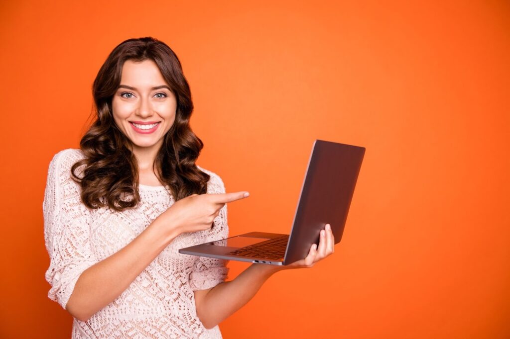 A smiling young woman with wavy brown hair holds an open laptop and points toward the screen against a solid orange background.