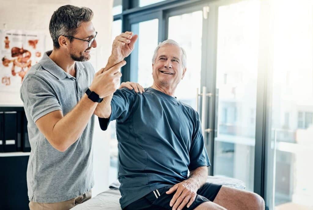 A male physical therapist gently guiding the arm of a senior man through a range-of-motion exercise.