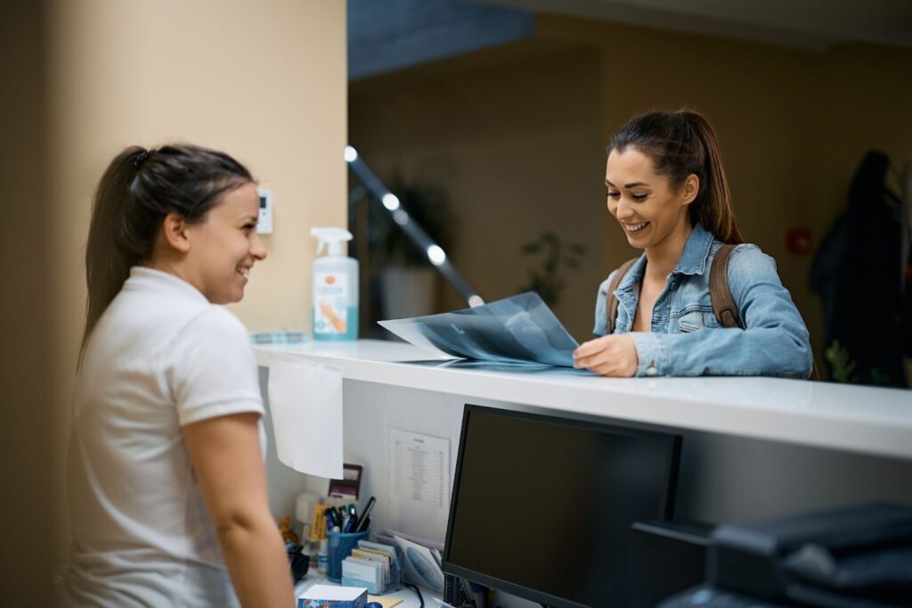 A smiling woman in a denim jacket stands at a clinic reception desk.