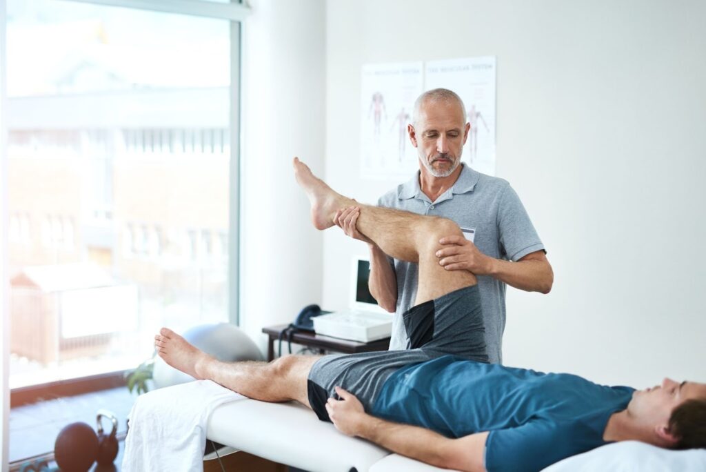 A physical therapist in a grey polo shirt performs a leg stretch and knee assessment on a male patient lying on a treatment table in a bright clinic.