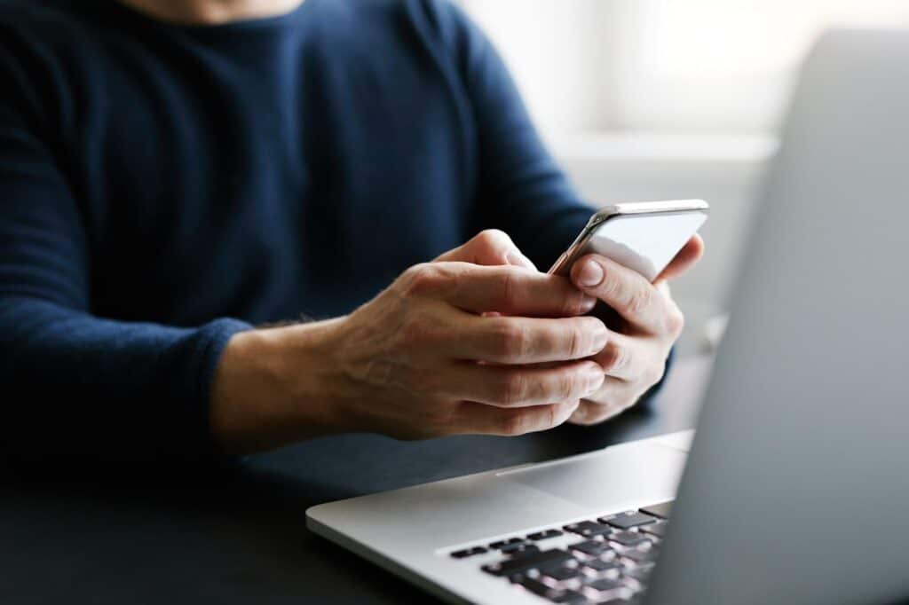 Close-up of a person's hands holding and using a smartphone while sitting at a desk with an open laptop.
