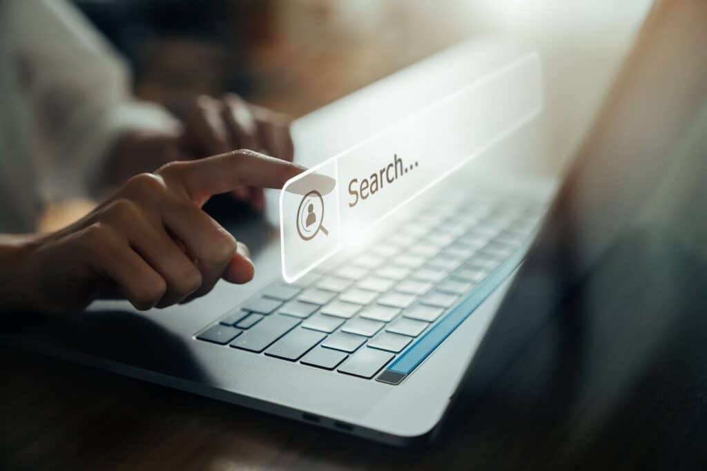 A close-up of a person's hand pressing a holographic, glowing search bar that floats above a laptop keyboard.
