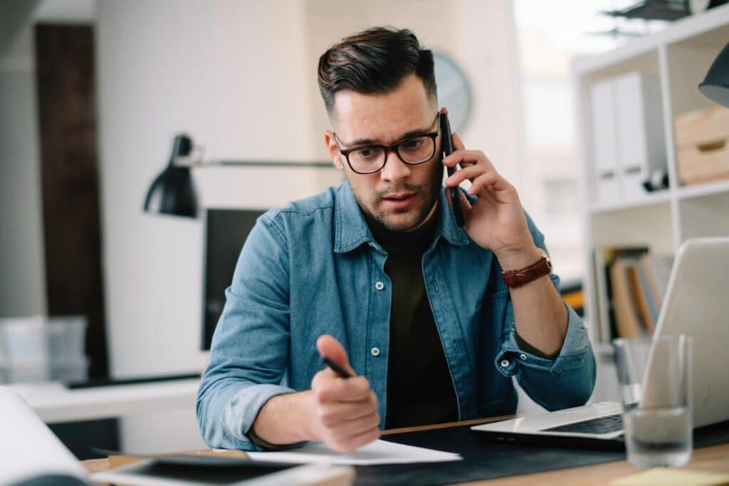 A man with glasses and a denim shirt, talking on a smartphone to schedule his PT appointment.