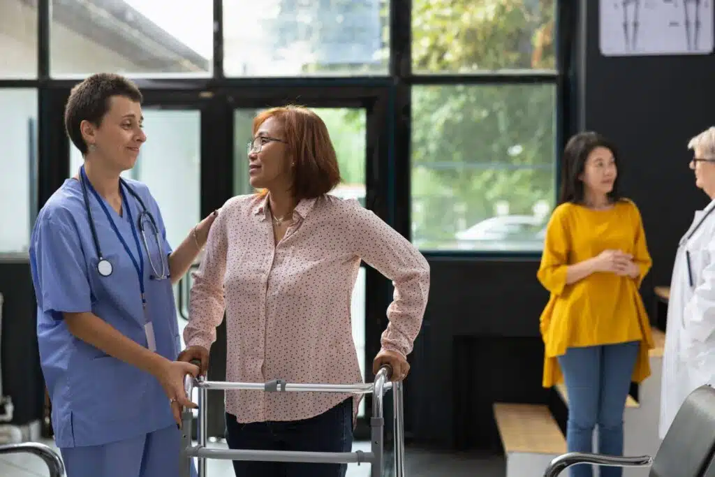 A healthcare professional in blue scrubs assists a woman using a walker in a bright medical facility.