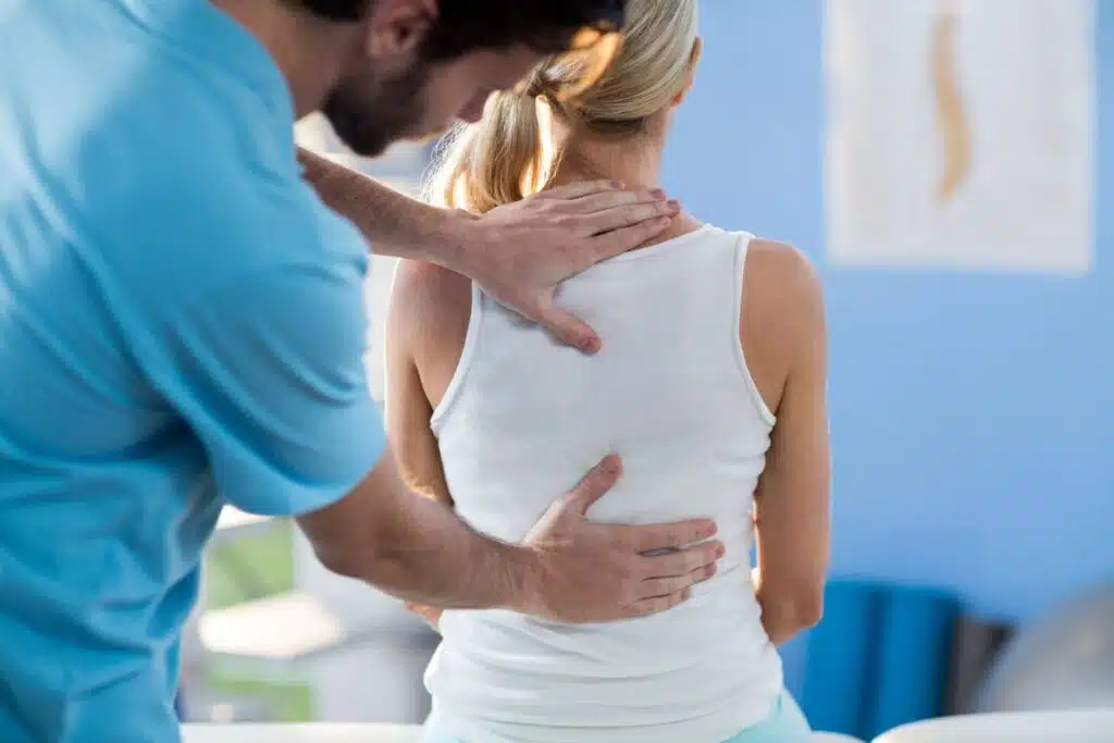 A male physical therapist in a blue scrub top performs a back assessment or treatment on a seated female patient.