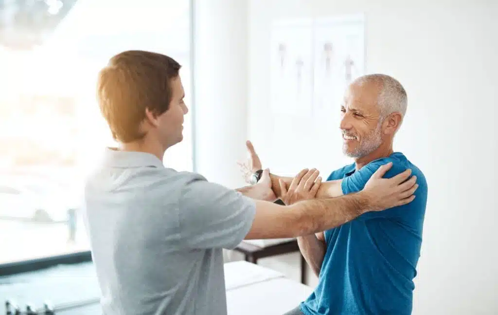 A physical therapist helps a man perform a shoulder stretch in a bright clinical setting. Both men are smiling during the session.